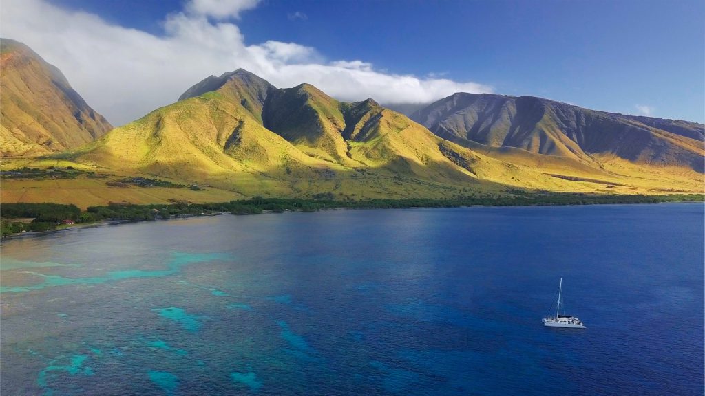 Sailboat on blue ocean near green mountains under a blue sky.