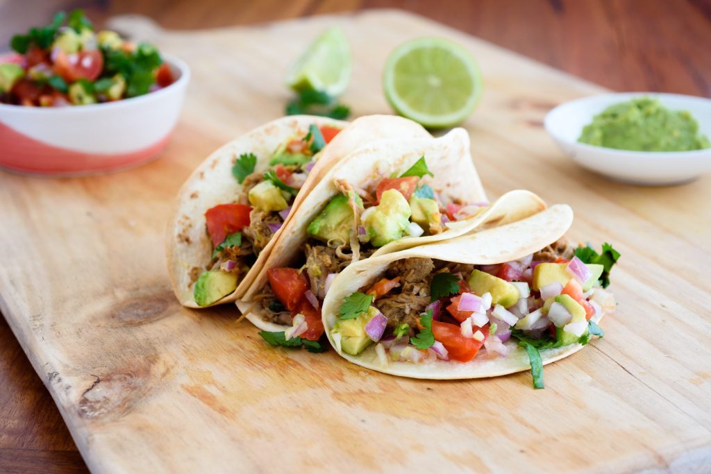 Two tacos with meat, avocado, and veggies on a wooden board, with lime and salsa on the side.