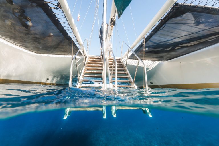 Partially submerged view beneath a catamaran with steps leading up, sails visible above water.