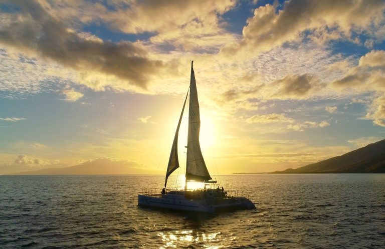 Silhouetted sailboat on ocean at sunset, with colorful sky and distant mountains.