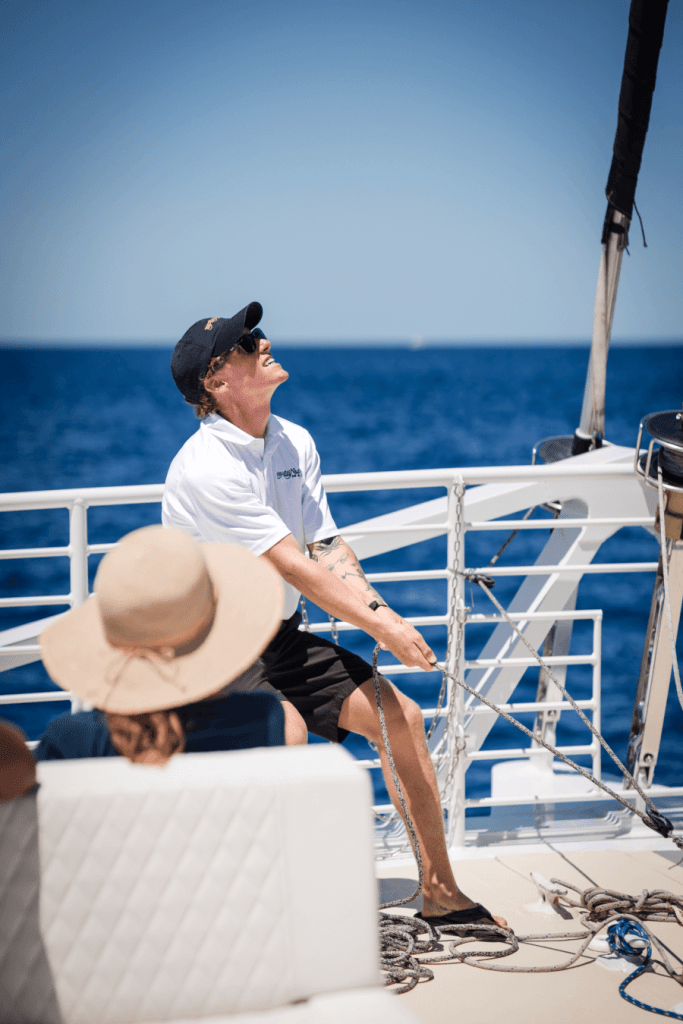 Two people on a boat deck, one adjusting sails, under a clear blue sky.