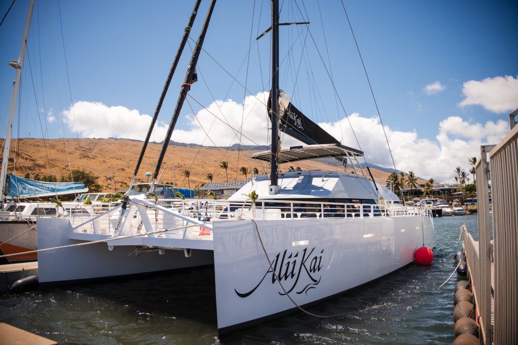White catamaran named Alii Kai docked by a pier under a clear sky.