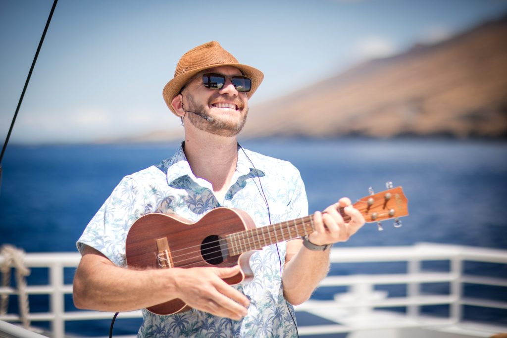 Man in hat and sunglasses playing ukulele on a boat.