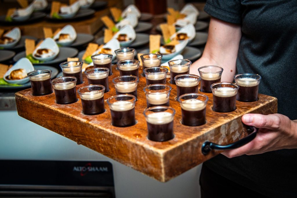 Person holding a tray of small glass cups filled with a creamy beverage.