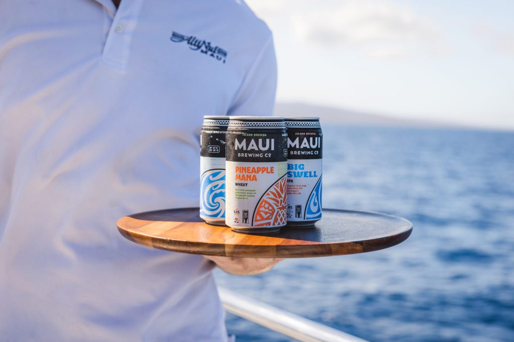Person holding a tray with Maui Brewing Co. cans by the ocean.