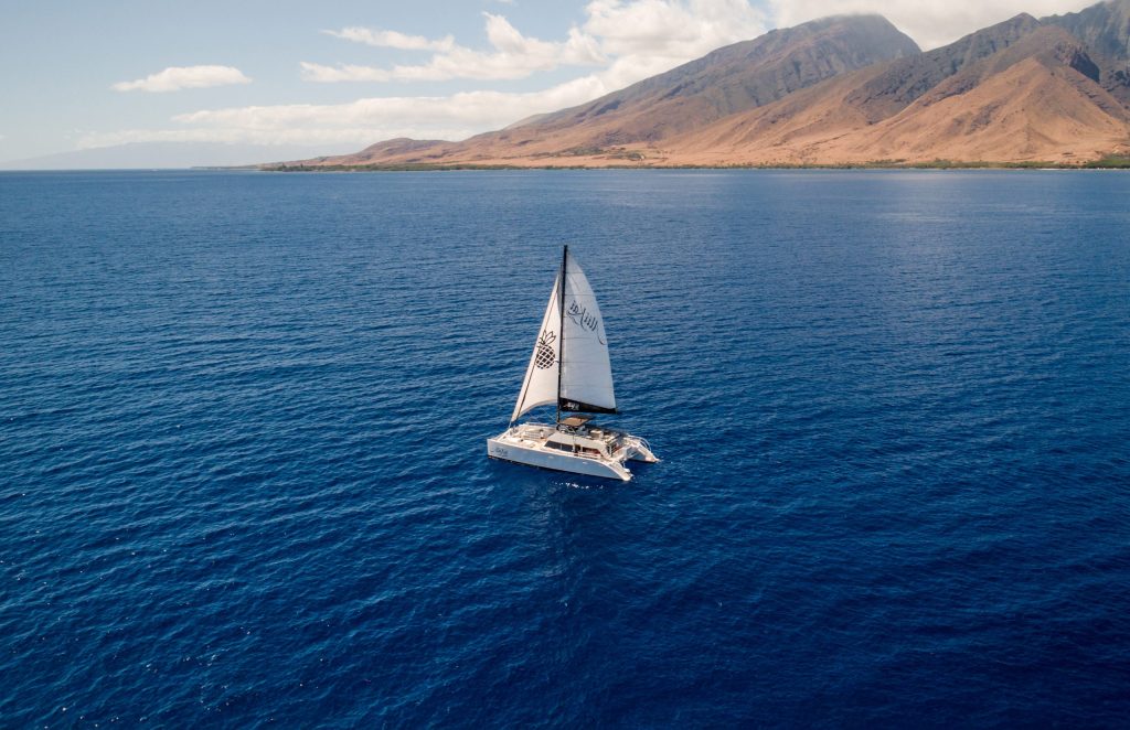 A catamaran sailing on a vast blue ocean near a mountainous coastline.