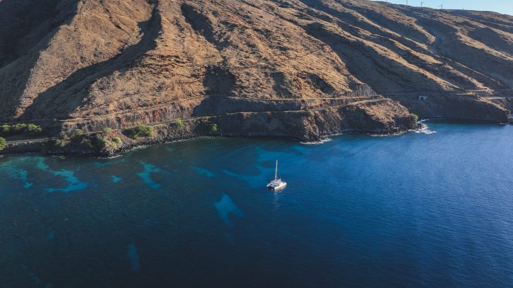 Sailboat on blue sea near rugged, dry hillside under a clear sky.