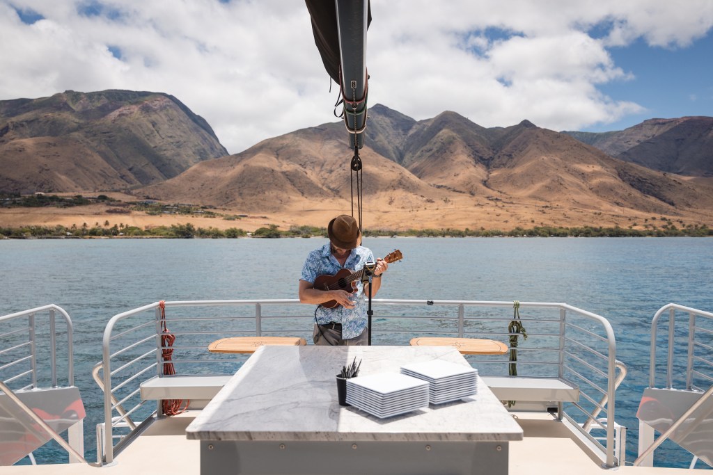Person playing ukulele on a boat with mountainous background.