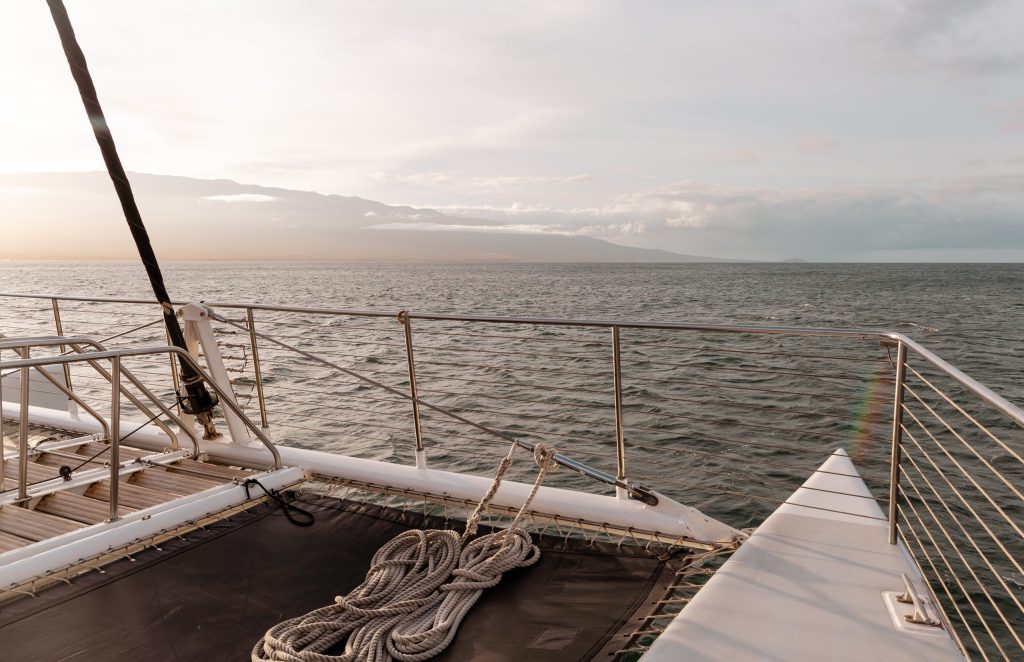 Sailboat deck with ropes and railings facing open sea under a cloudy sky.
