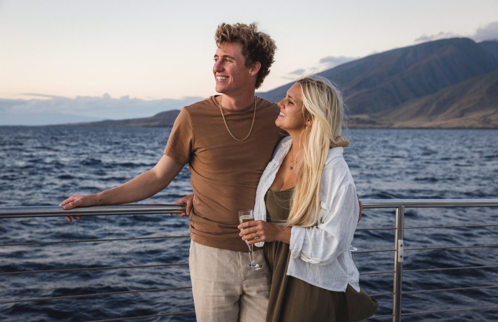 Couple smiling on a boat, gazing at the ocean with mountains in the background.