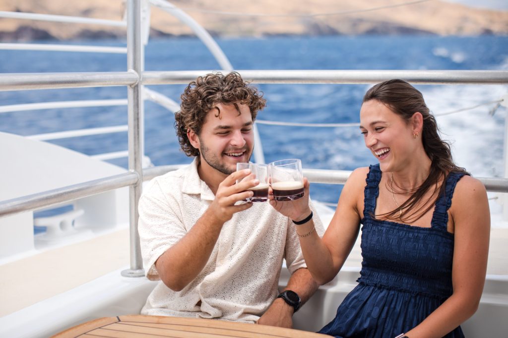 A smiling couple toasting with drinks on a boat overlooking the ocean.