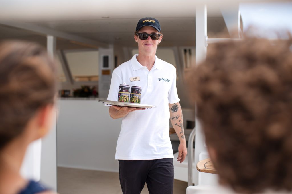 Smiling person with a tray of drinks on a boat, wearing a white polo and sunglasses.