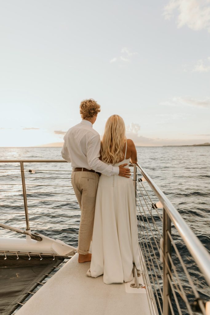 Couple in formal wear standing on yacht, looking at ocean sunset.
