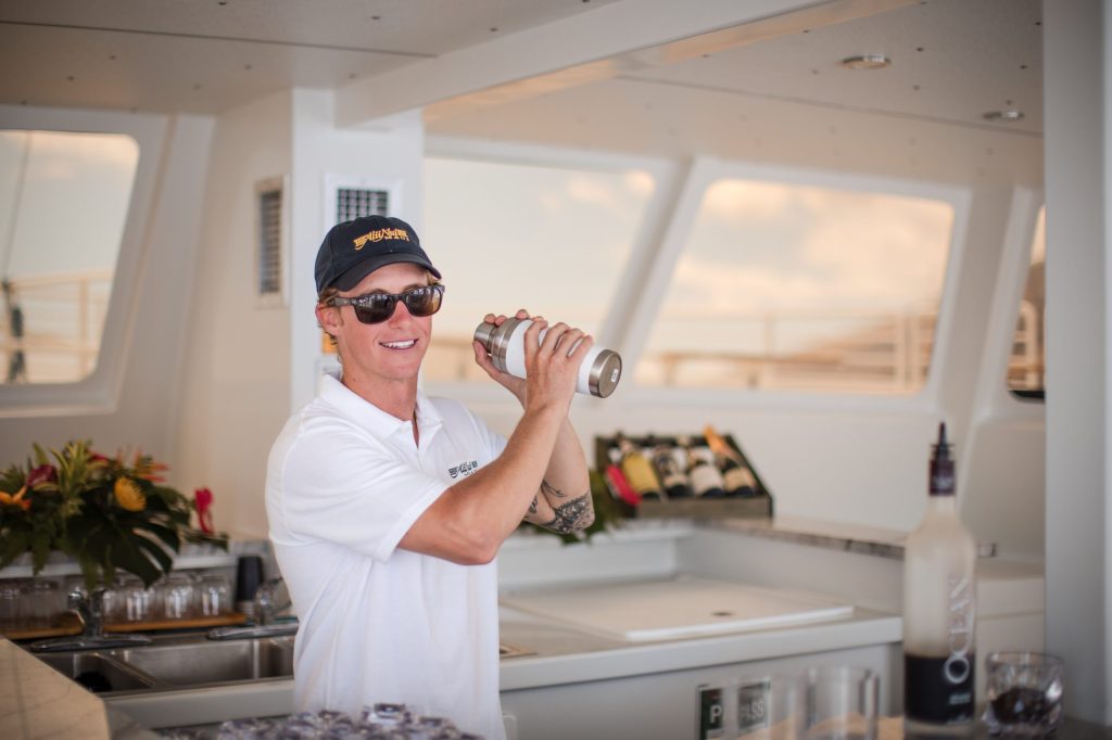 Person in a cap and sunglasses shaking a cocktail shaker on a boat.