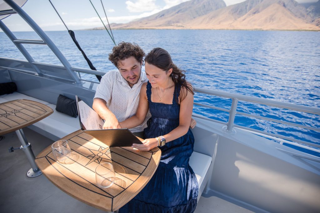 Couple on a boat looking at a laptop with ocean and mountains in the background.