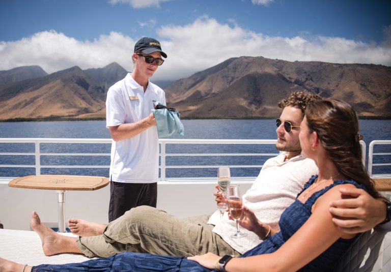 Couple relaxing on a yacht, served by a staff member, with scenic mountains in the background.
