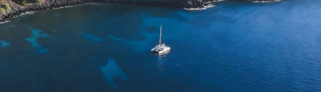 Aerial view of a sailboat on a vast blue ocean near a rocky coastline.