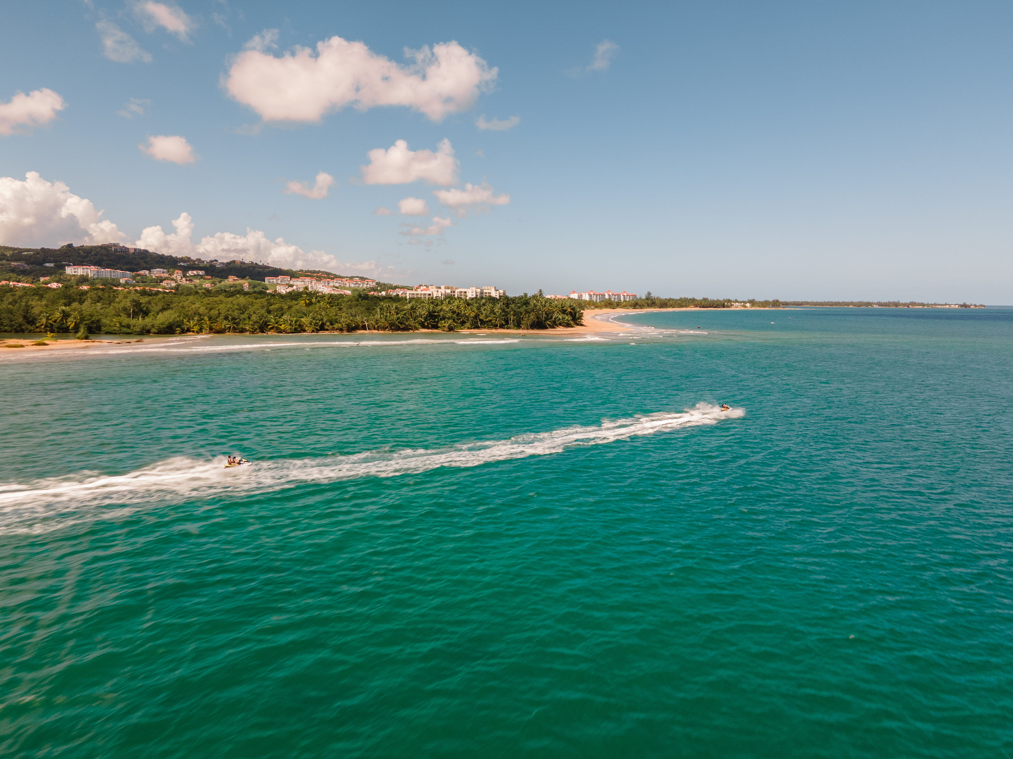 Jet skis leaving white trails on turquoise water near a green coastline.