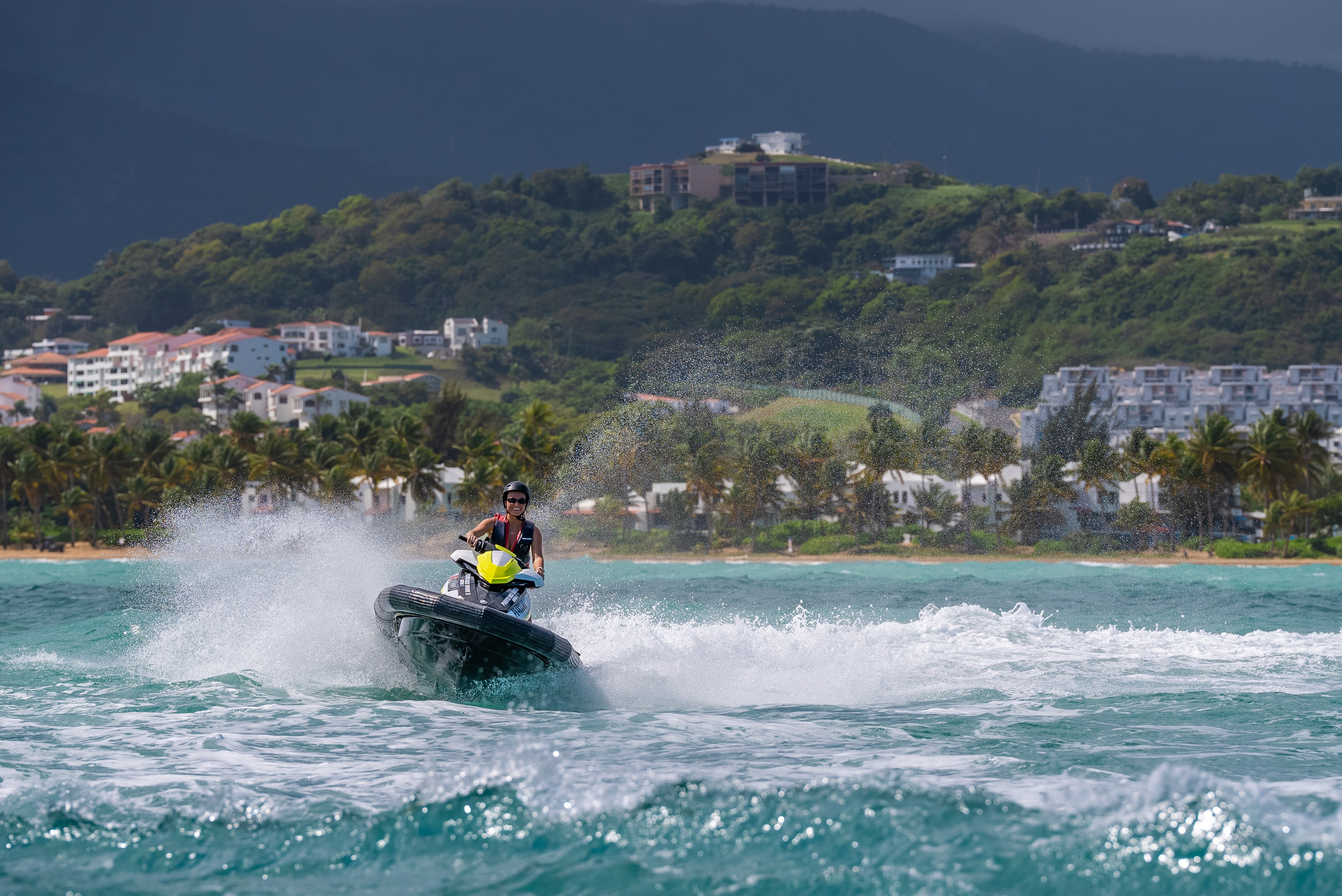 Person riding a jet ski on turquoise water with tropical shore and mountains in the background.