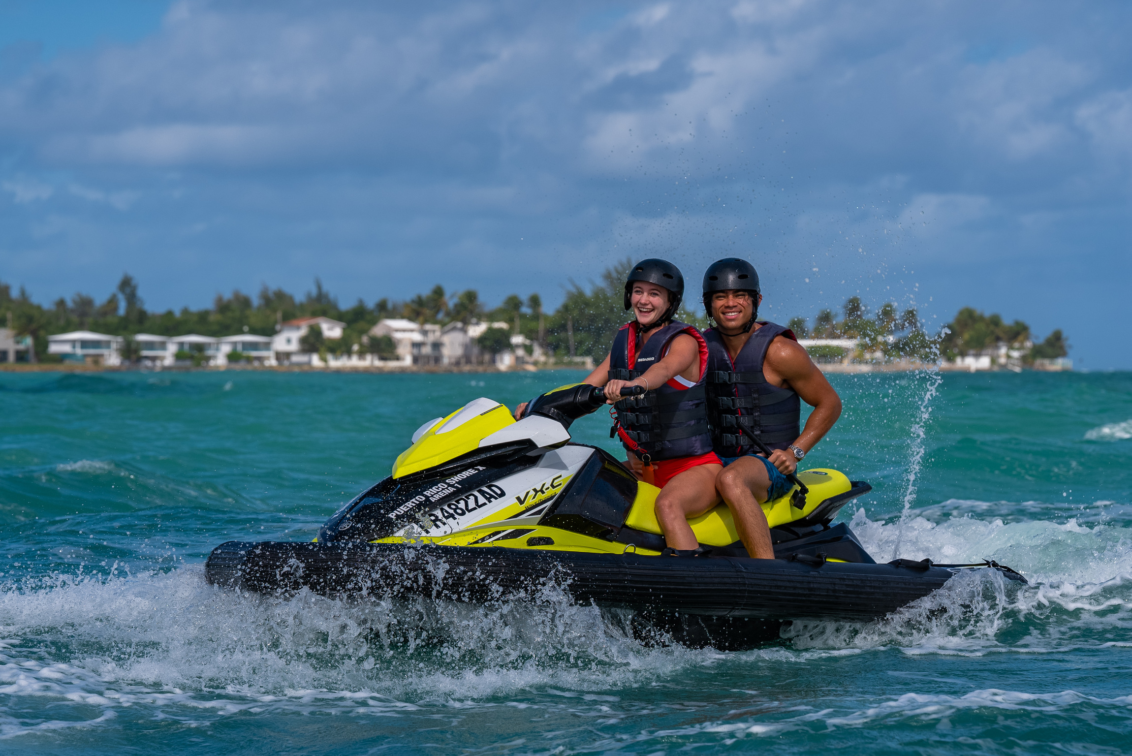 Smiling couple riding a yellow jet ski on the ocean with buildings in the background.