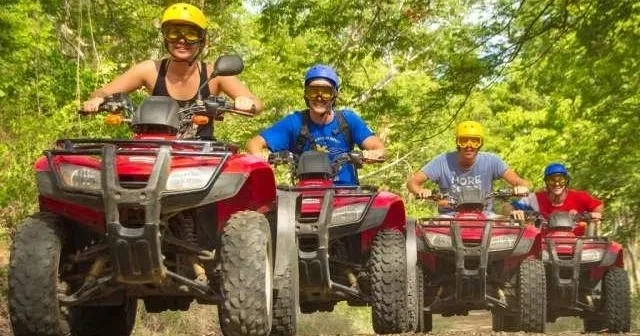 Group of four people riding red ATVs through a forest trail.