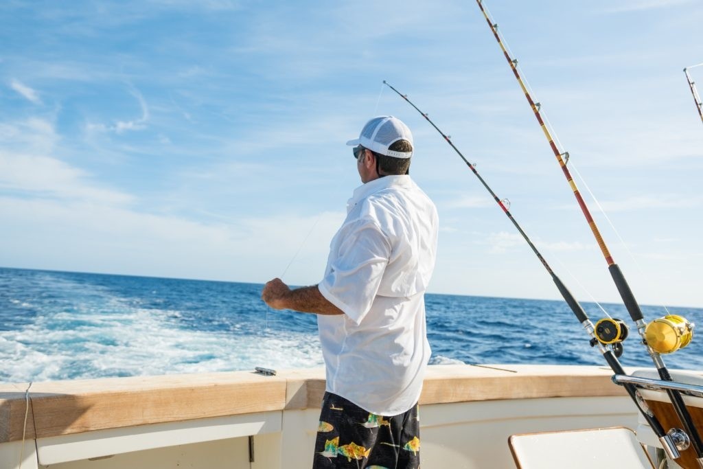 Man fishing on a boat in open ocean, wearing white shirt and cap, with fishing rods visible.
