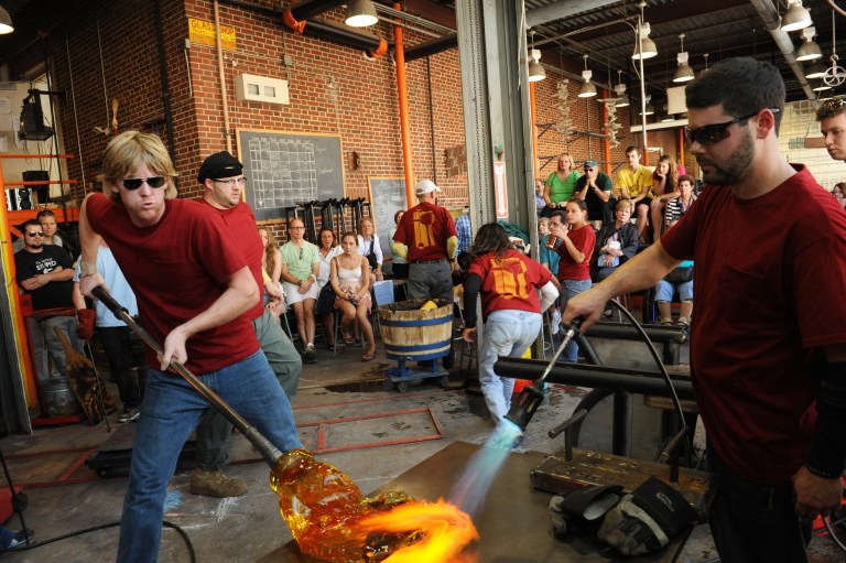 Glassblowers in red shirts work with molten glass in front of an audience.