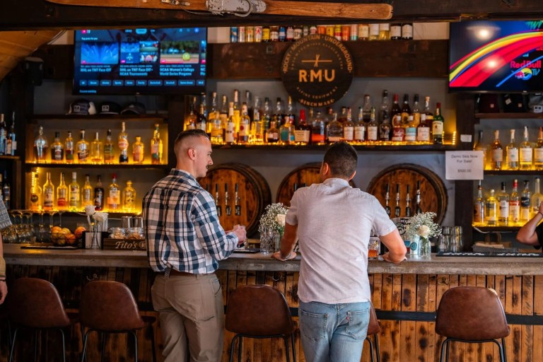 Two men at a rustic bar with bottles and screens in the background.