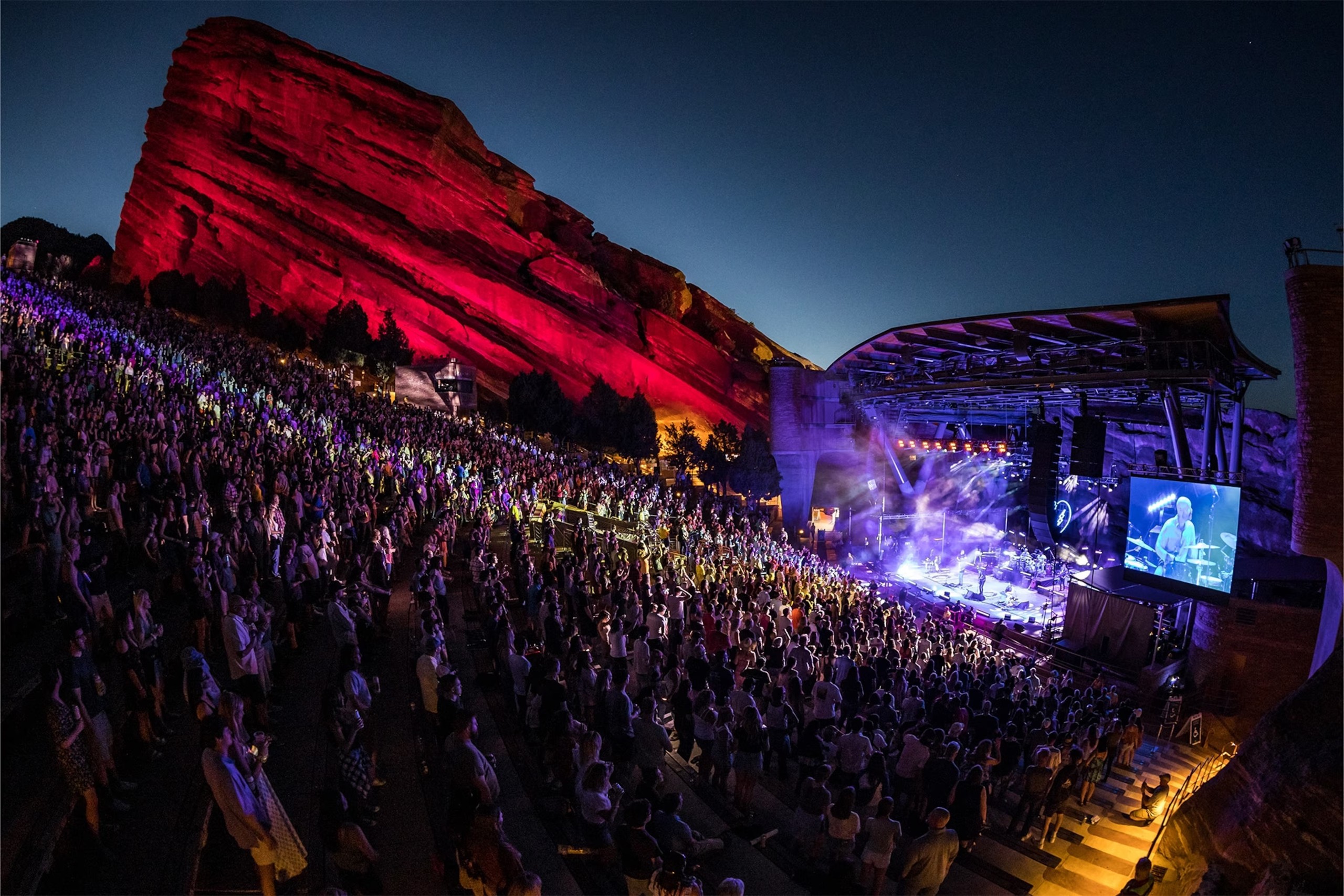 Night concert at Red Rocks Amphitheatre, audience facing lit stage and red illuminated rock formation.