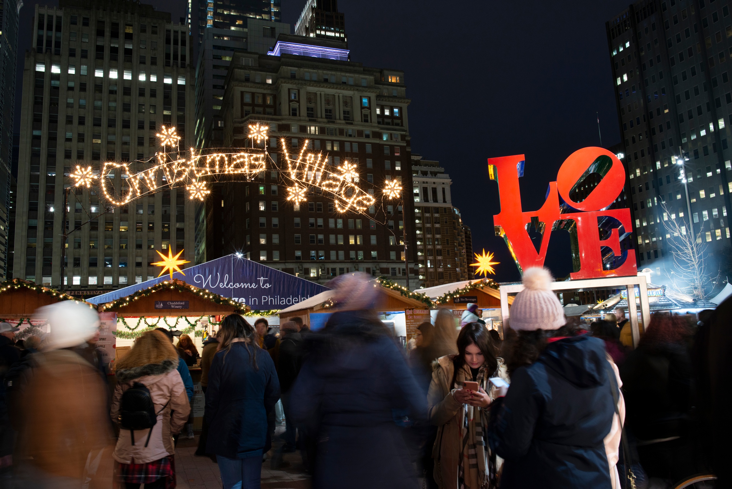 Crowded Christmas market at night in city with bright 'Christmas Village' sign.
