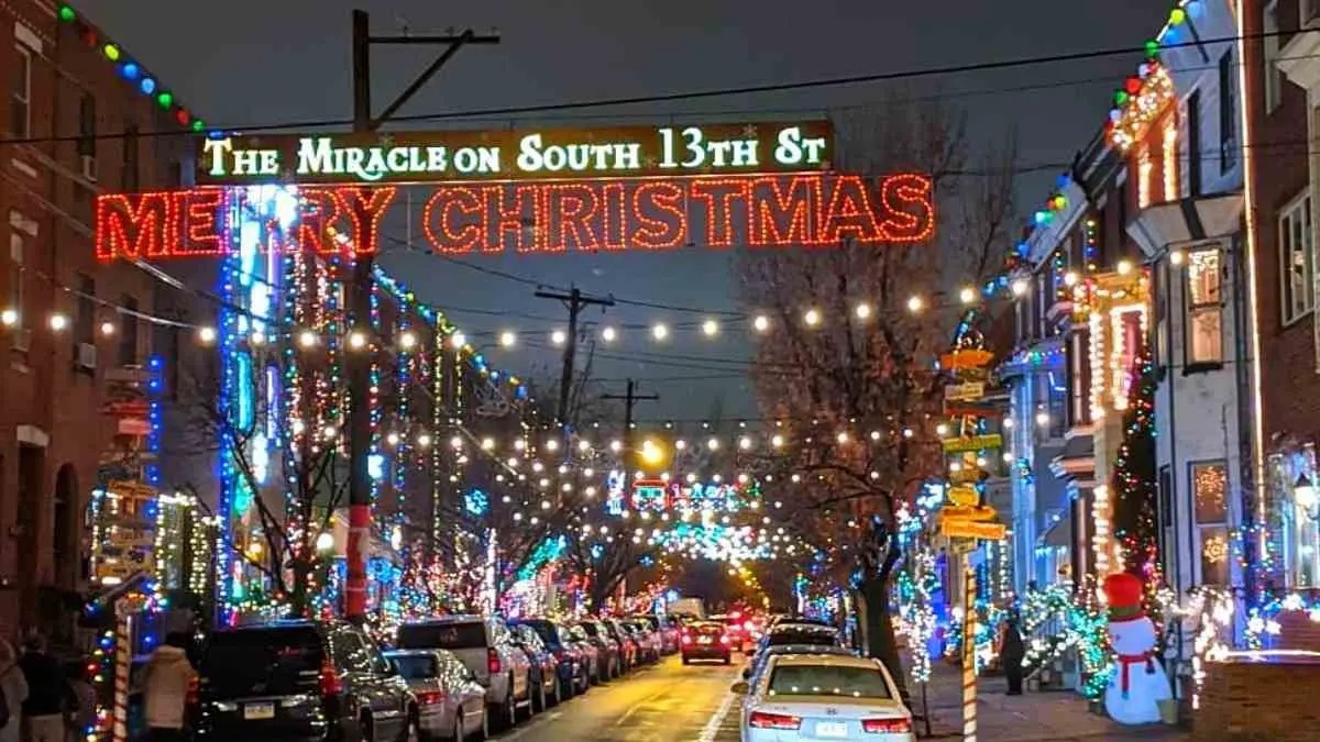 Festively lit street with 'Merry Christmas' sign and houses decorated with lights.