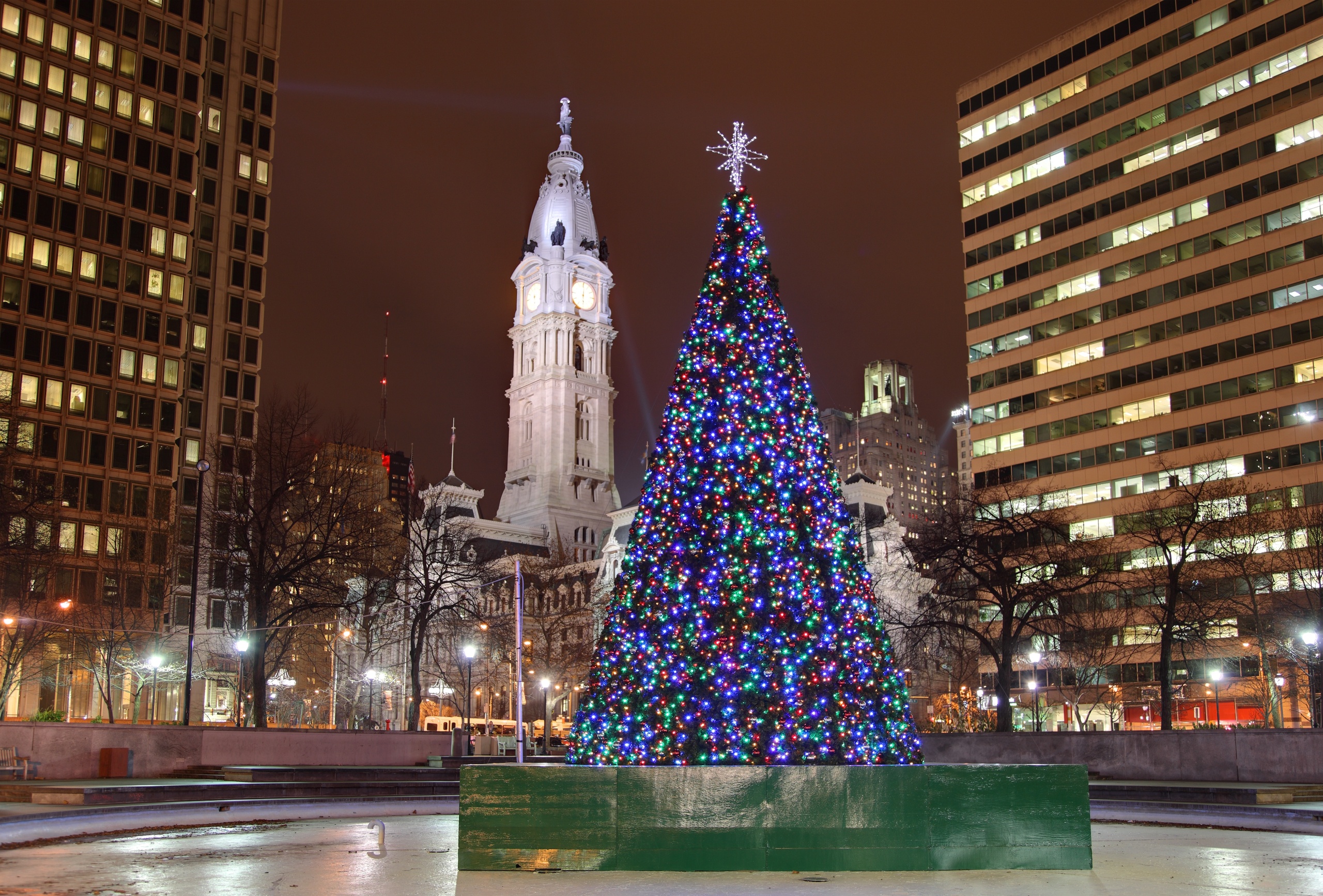 Colorful Christmas tree in plaza with clock tower and tall buildings in background, nighttime.