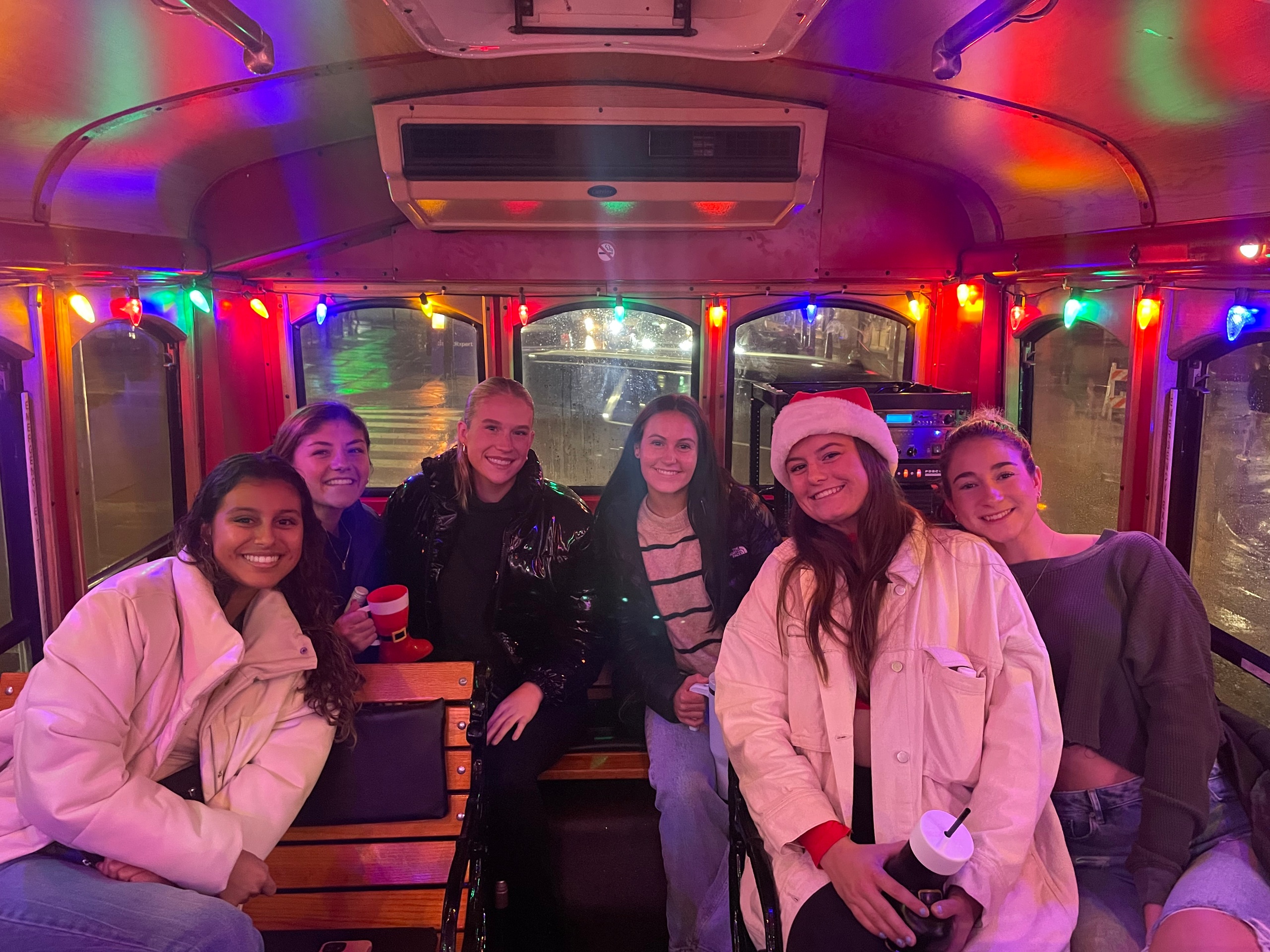 Six people smiling inside a festively lit trolley with colorful lights.