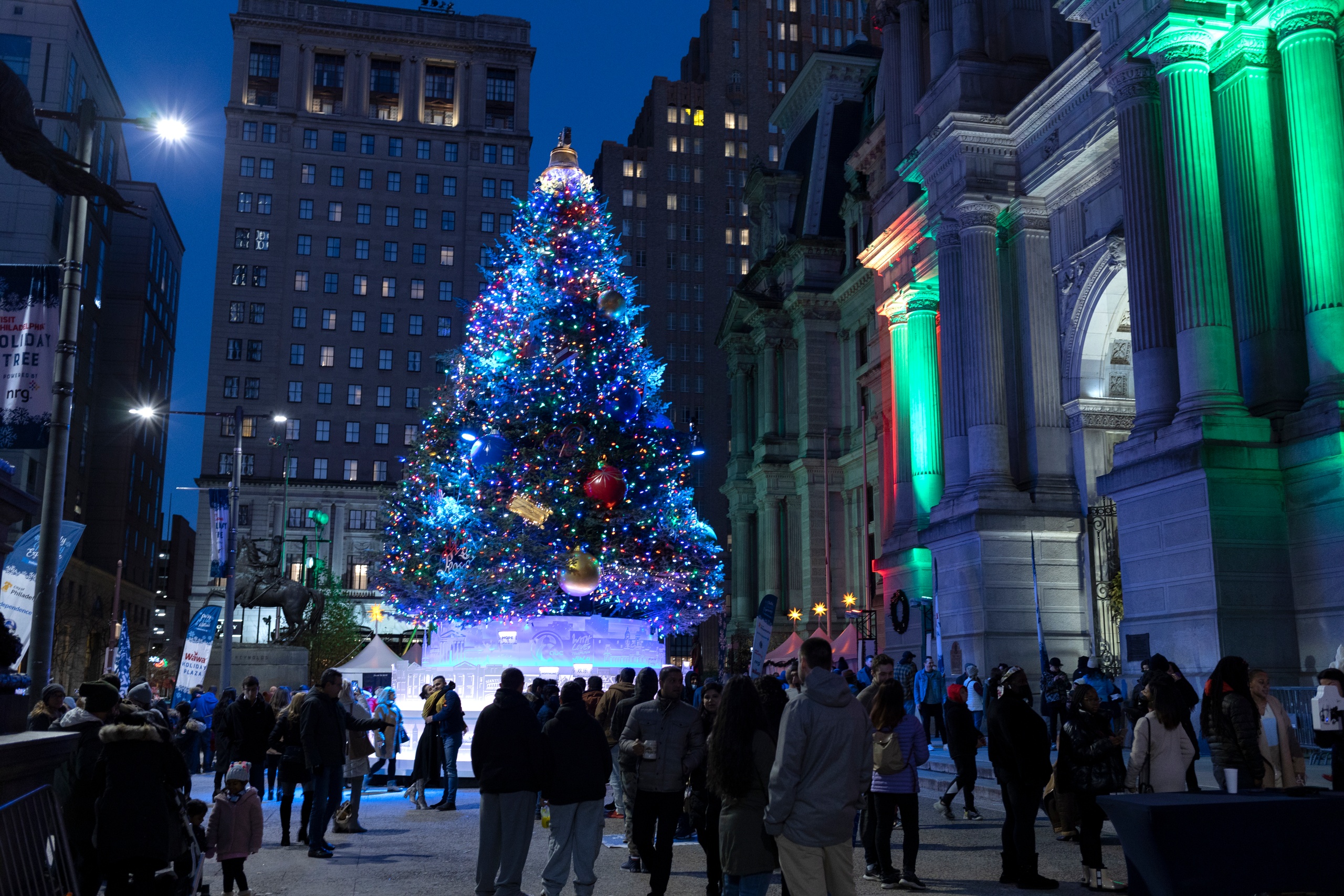 Crowd gathers around a large, decorated Christmas tree in a city plaza at night.