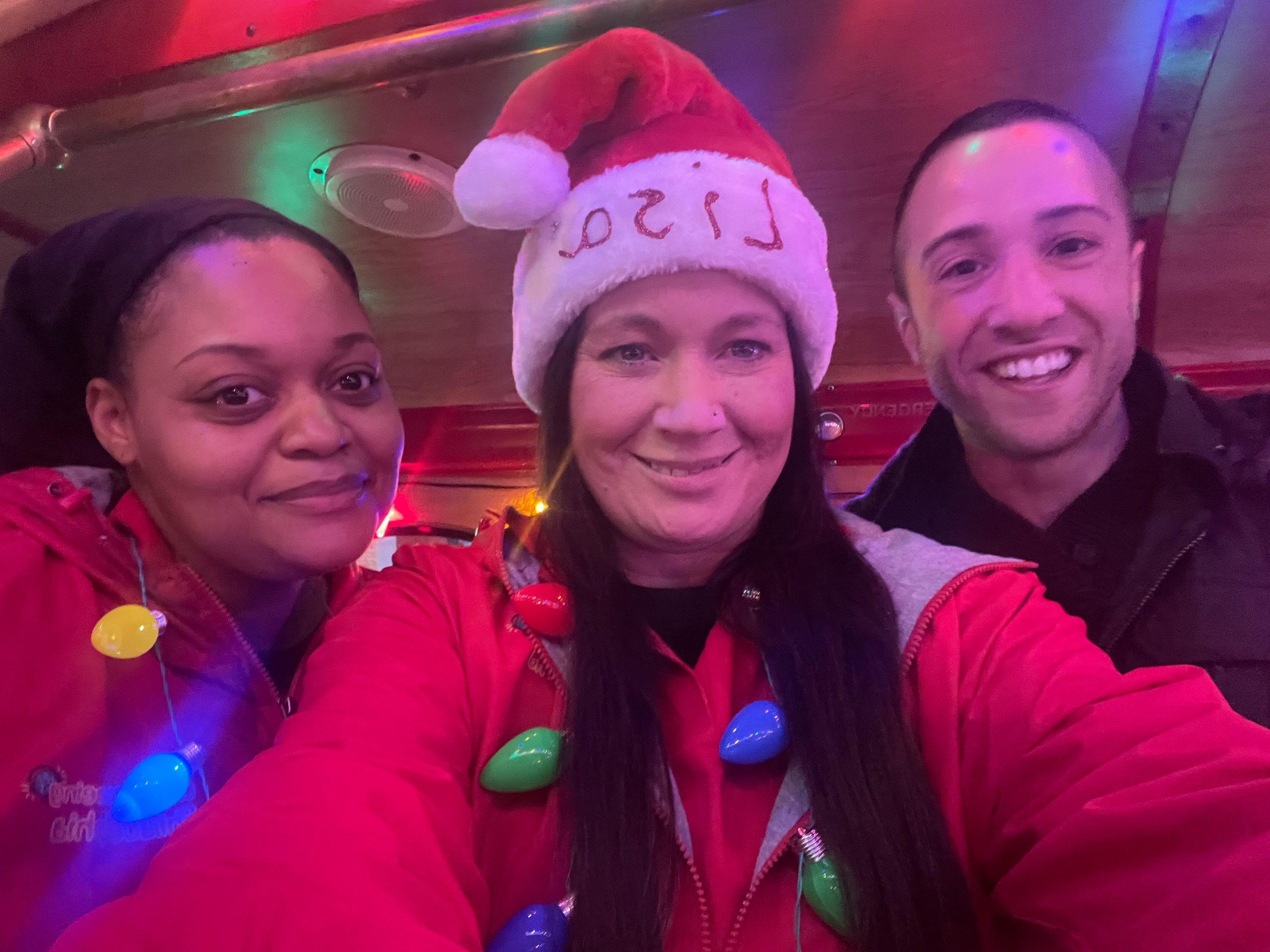 Three people smiling, one wearing a Santa hat and colorful lights necklace.