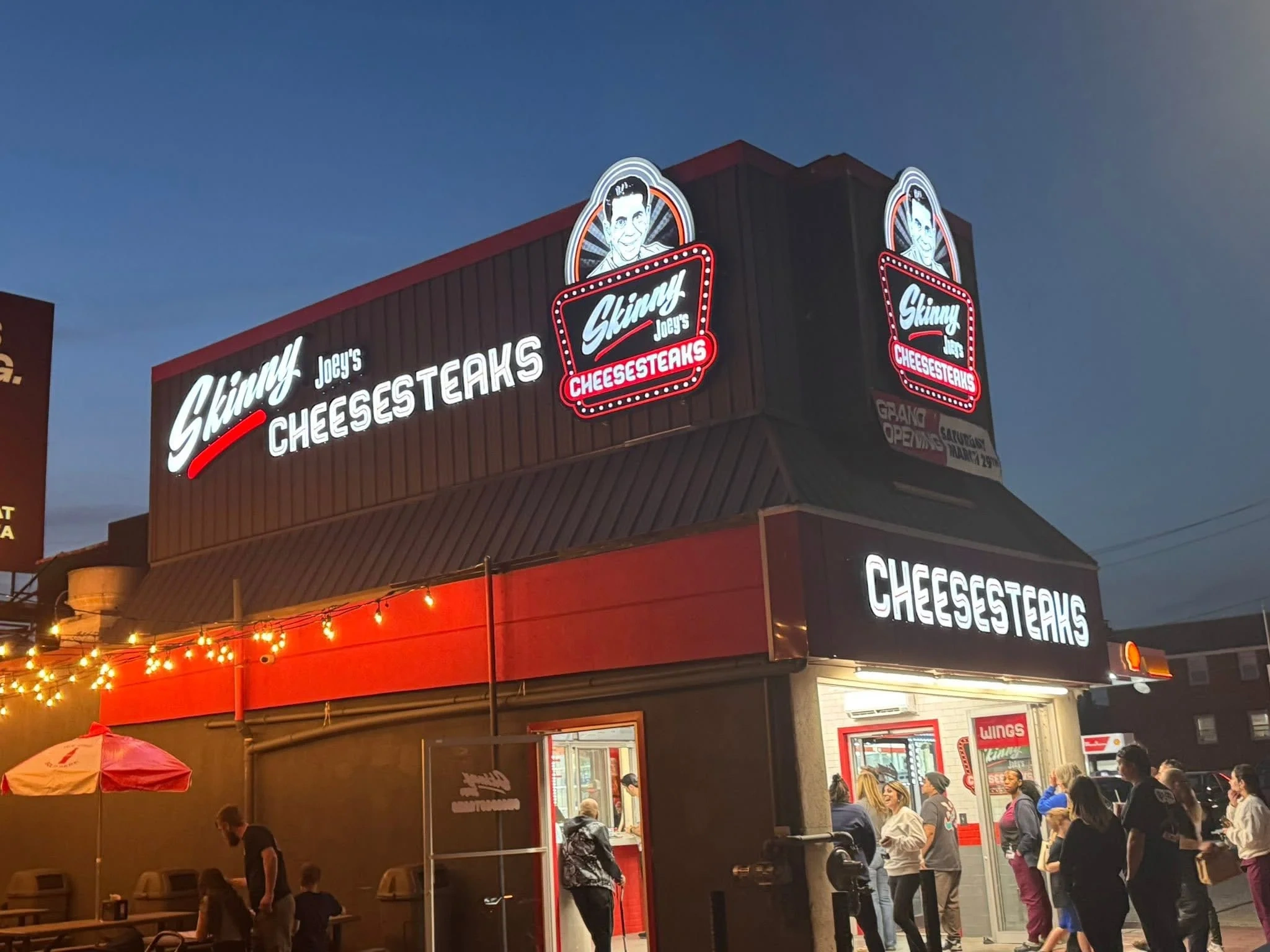 Illuminated cheesesteak restaurant at dusk with customers and string lights.