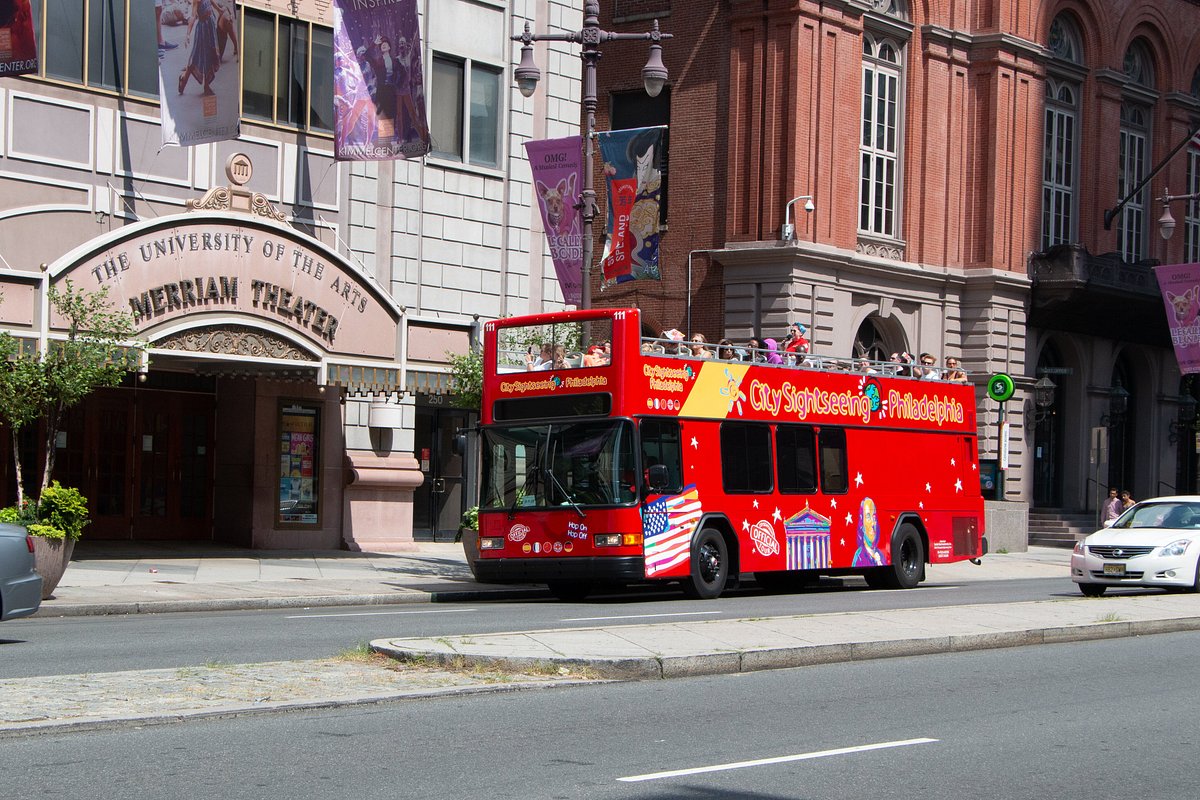 Red double-decker tour bus parked near Merriam Theater on city street.