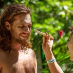 Person applying face paint to a smiling man outdoors in a lush area.