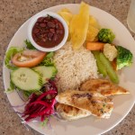 Plate of grilled chicken, rice, beans, plantains, and mixed vegetables on a table.