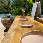 Wooden table with empty plates, bamboo cup, and menu near a pool and lush greenery.
