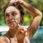 Woman applying face mask with brush outdoors in a garden setting.