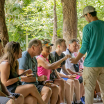 People seated outdoors interacting with a standing person in a green shirt, surrounded by trees.