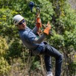 Person zip-lining in forest, wearing helmet and gloves, giving thumbs-up.