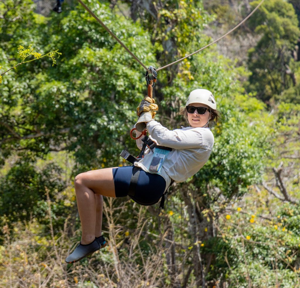 Person ziplining through a lush green forest wearing sunglasses and a helmet.