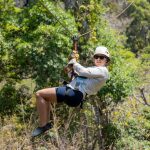 Person ziplining through a lush green forest wearing sunglasses and a helmet.