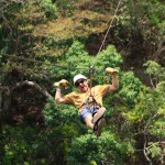 Person zip-lining through a forest wearing a helmet and gloves, surrounded by greenery.