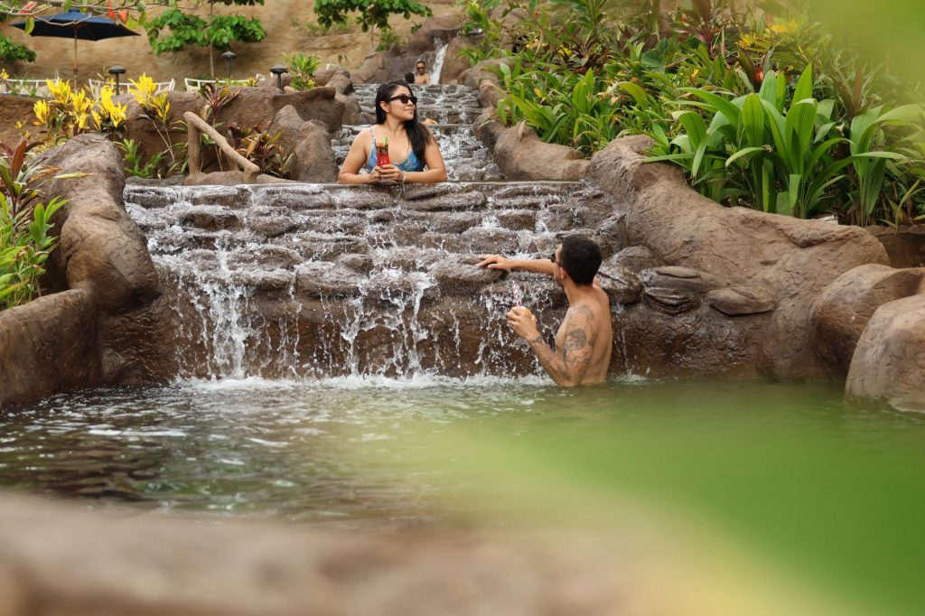 Two people enjoying a natural hot spring waterfall surrounded by greenery.