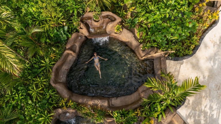Person floating in a circular natural pool surrounded by lush greenery.