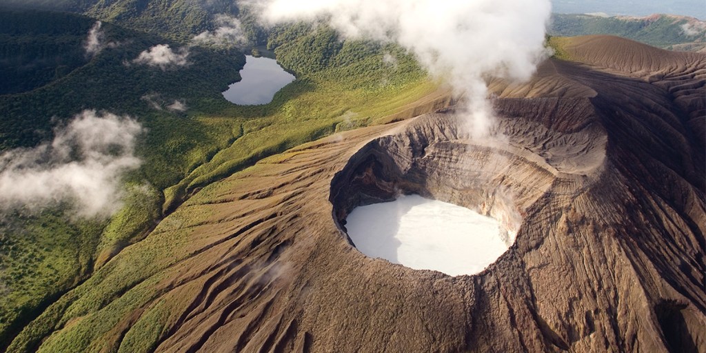 Aerial view of a smoking volcano crater surrounded by green forest and clouds.