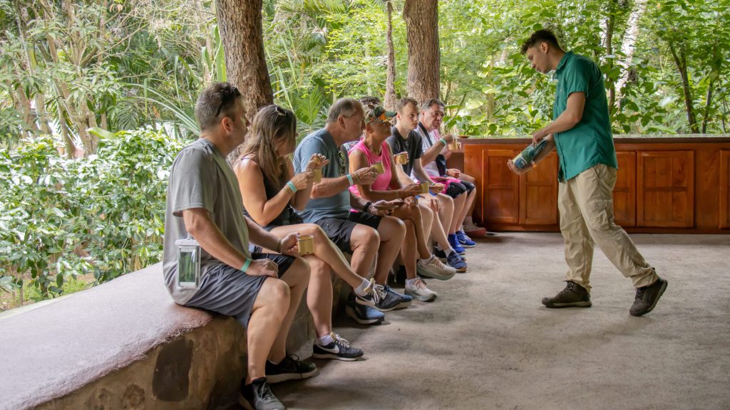 Group of people sitting on a bench watching a man in a green shirt showing an object.